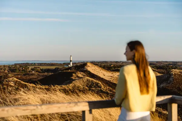 Eine Frau steht an einem Zaun und blickt auf einen Leuchtturm und Dünen.