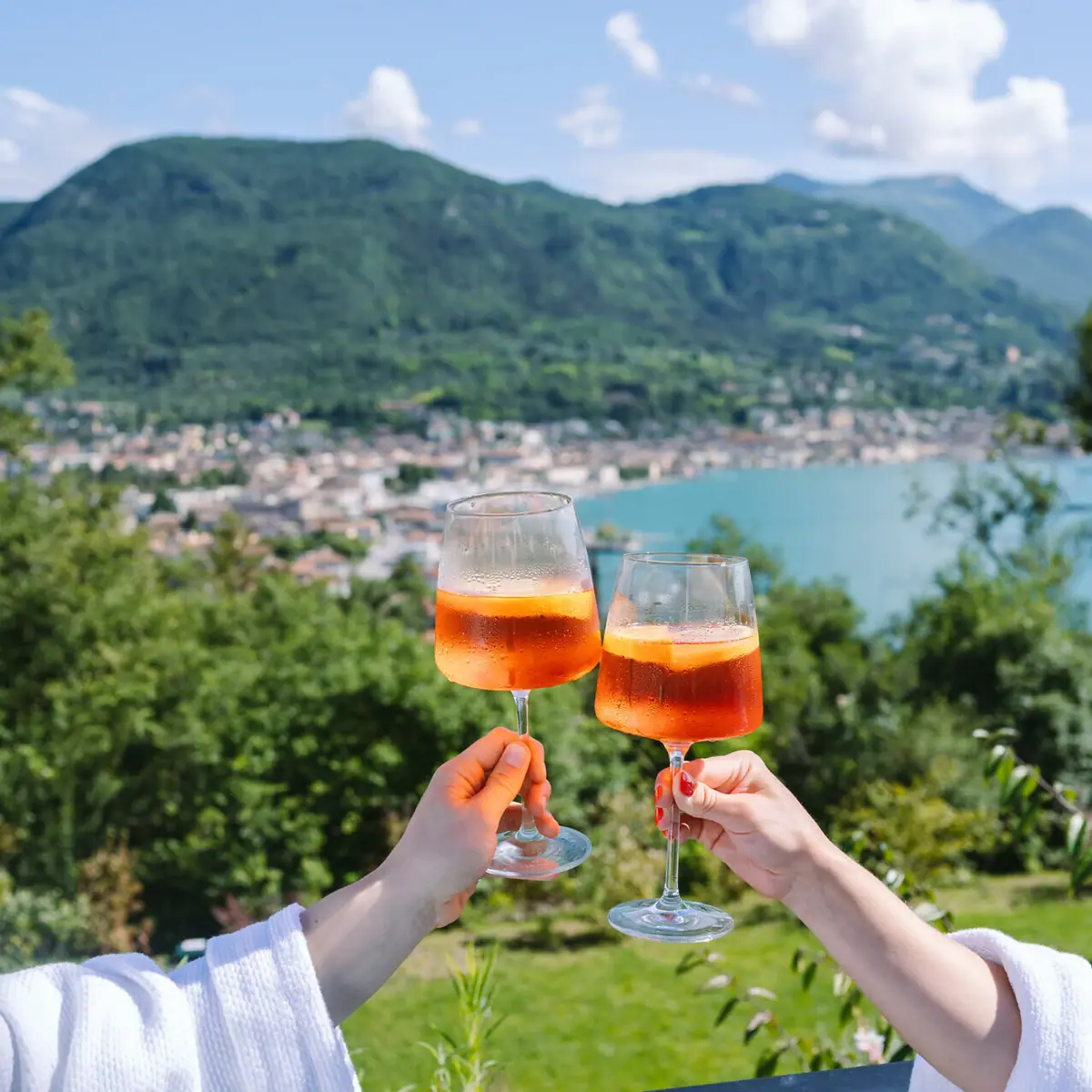Zwei Hände halten Weingläser mit Aperol Spritz vor dem Gardasee.