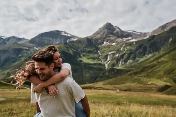 Ein Mann trägt eine Frau auf dem Rücken in einem grasbewachsenen Feld mit Bergen im Hintergrund.