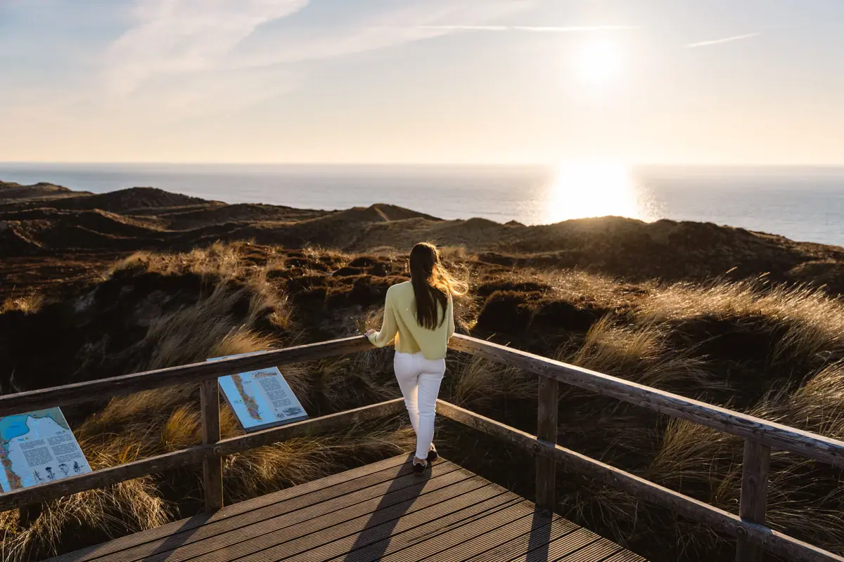 Eine Frau steht auf einer Holzbrücke und blickt auf die Wanderdünen und die Nordsee auf Sylt.